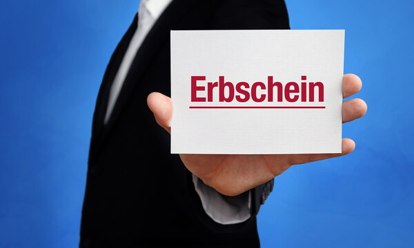 Ein weißes Schild mit dem Wort „Erbschein“ in roter Farbe wird in von einem Mann in Anzug in die Kamera gehalten.