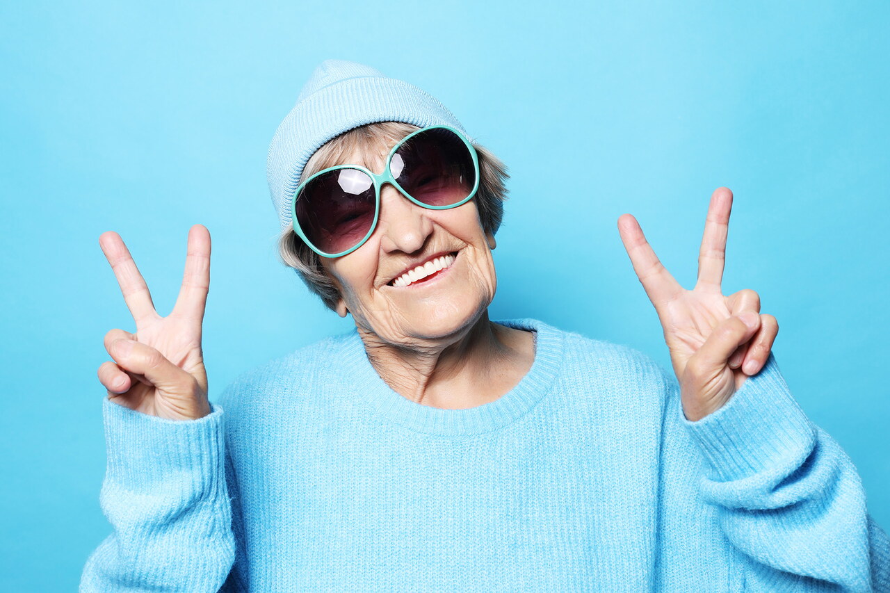Eine jung gebliebene Oma hat eine Mütze und eine Sonnenbrille an und zeigt mit beiden Händen das Peace-Zeichen in die Kamera.