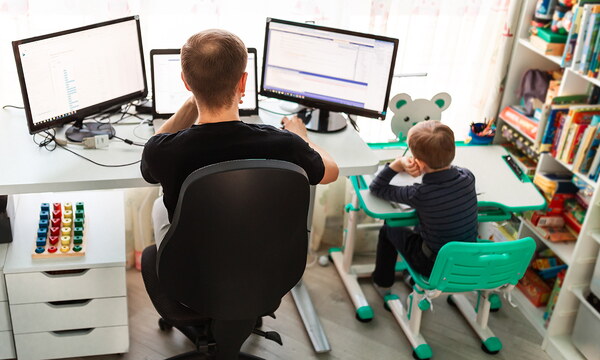 Ein Arbeitnehmer sitzt im Home Office an seinem Schreibtisch. Direkt neben ihm steht ein kleiner Schreibtisch, an dem ein Kind die Hausaufgaben macht.