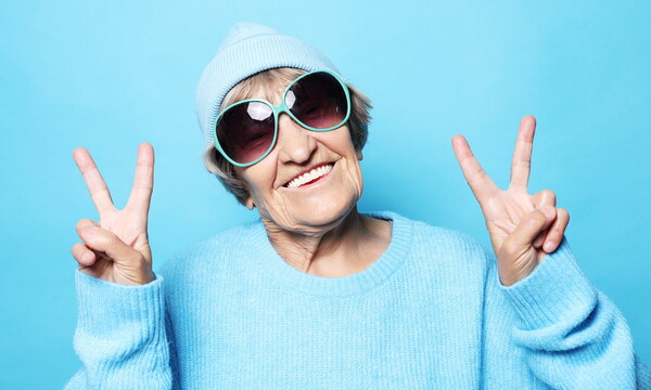 Eine jung gebliebene Oma hat eine Mütze und eine Sonnenbrille an und zeigt mit beiden Händen das Peace-Zeichen in die Kamera.