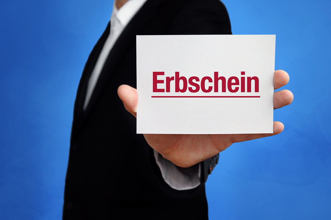 Ein weißes Schild mit dem Wort „Erbschein“ in roter Farbe wird in von einem Mann in Anzug in die Kamera gehalten.