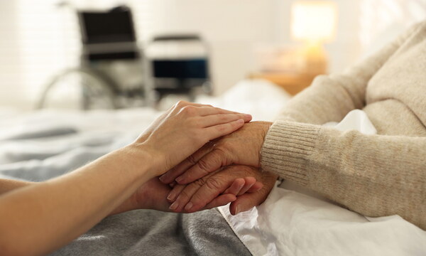 EIne Seniorin liegt in einem Bett, ihre Hände werden von einer jüngeren Person gehalten. Im Hintergrund steht ein Rollstuhl.