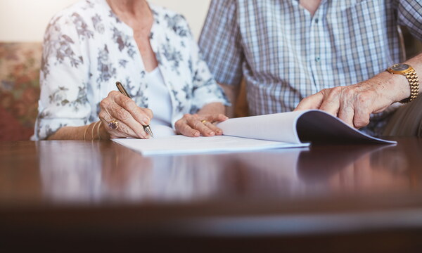 Ein älteres Paar sitzt vor einem Dokument, sie unterschreibt gerade. Die Gesichter sind nicht zu sehen.