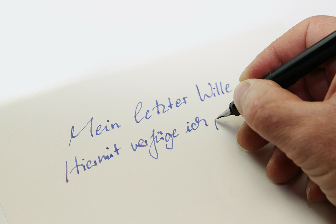 Jemand schreibt mit Füller auf ein Blatt Papier: „Mein letzter Wille. Hiermit verfüge ich, …“
