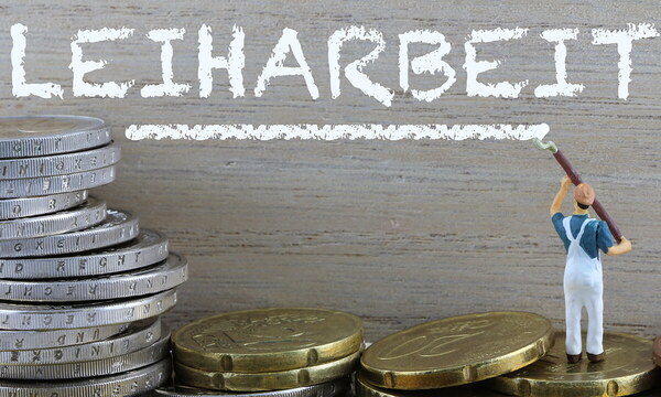 Nahaufnahme: Vor einer Wand sind Münzen gestapelt, während eine darauf stehende Spielzeugfigur das Wort Leiharbeit an die Wand pinselt.