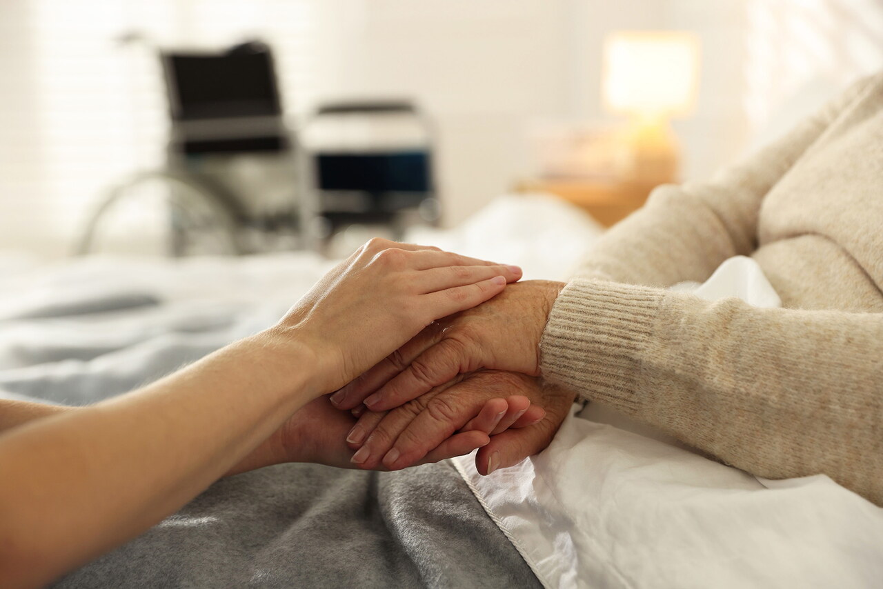 EIne Seniorin liegt in einem Bett, ihre Hände werden von einer jüngeren Person gehalten. Im Hintergrund steht ein Rollstuhl.