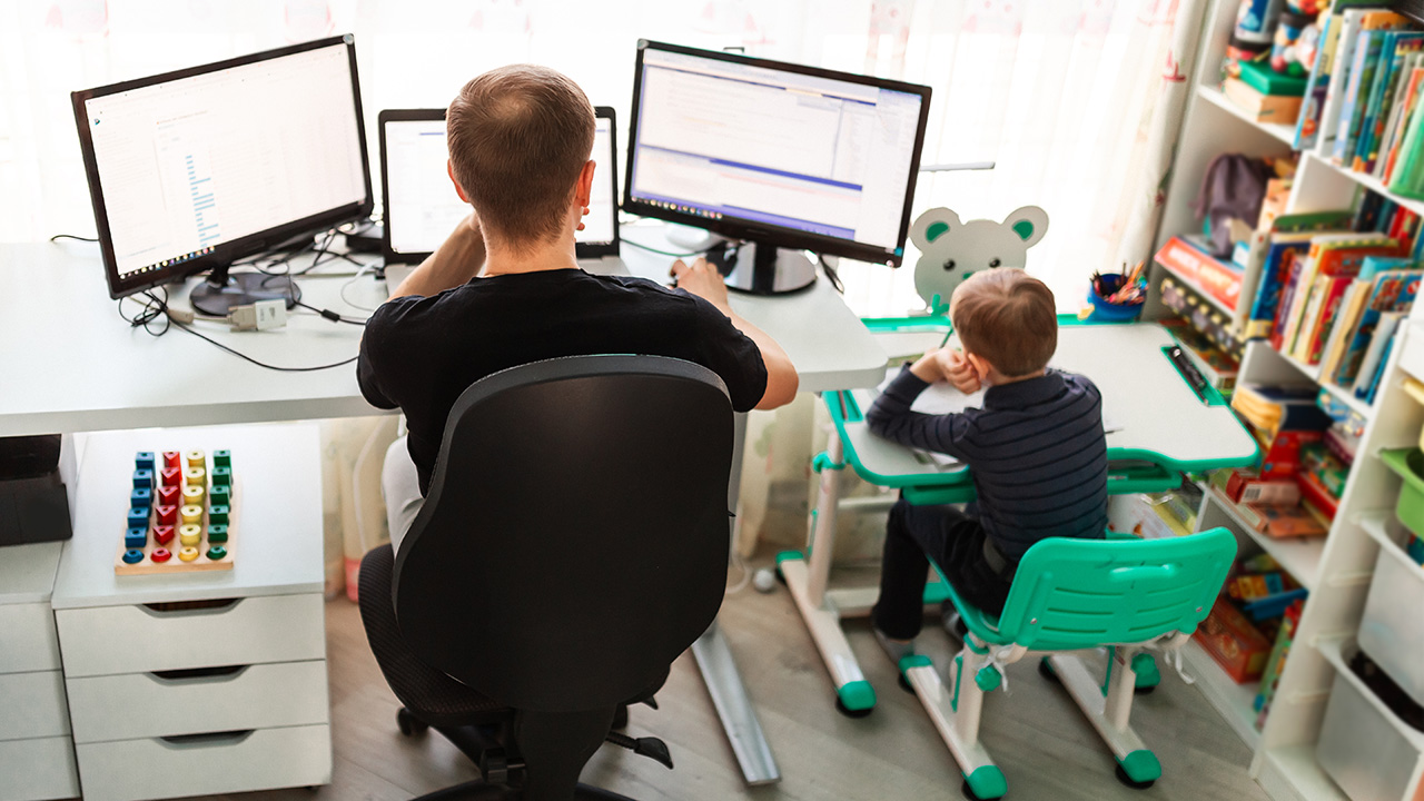 Ein Arbeitnehmer sitzt im Home Office an seinem Schreibtisch. Direkt neben ihm steht ein kleiner Schreibtisch, an dem ein Kind die Hausaufgaben macht.